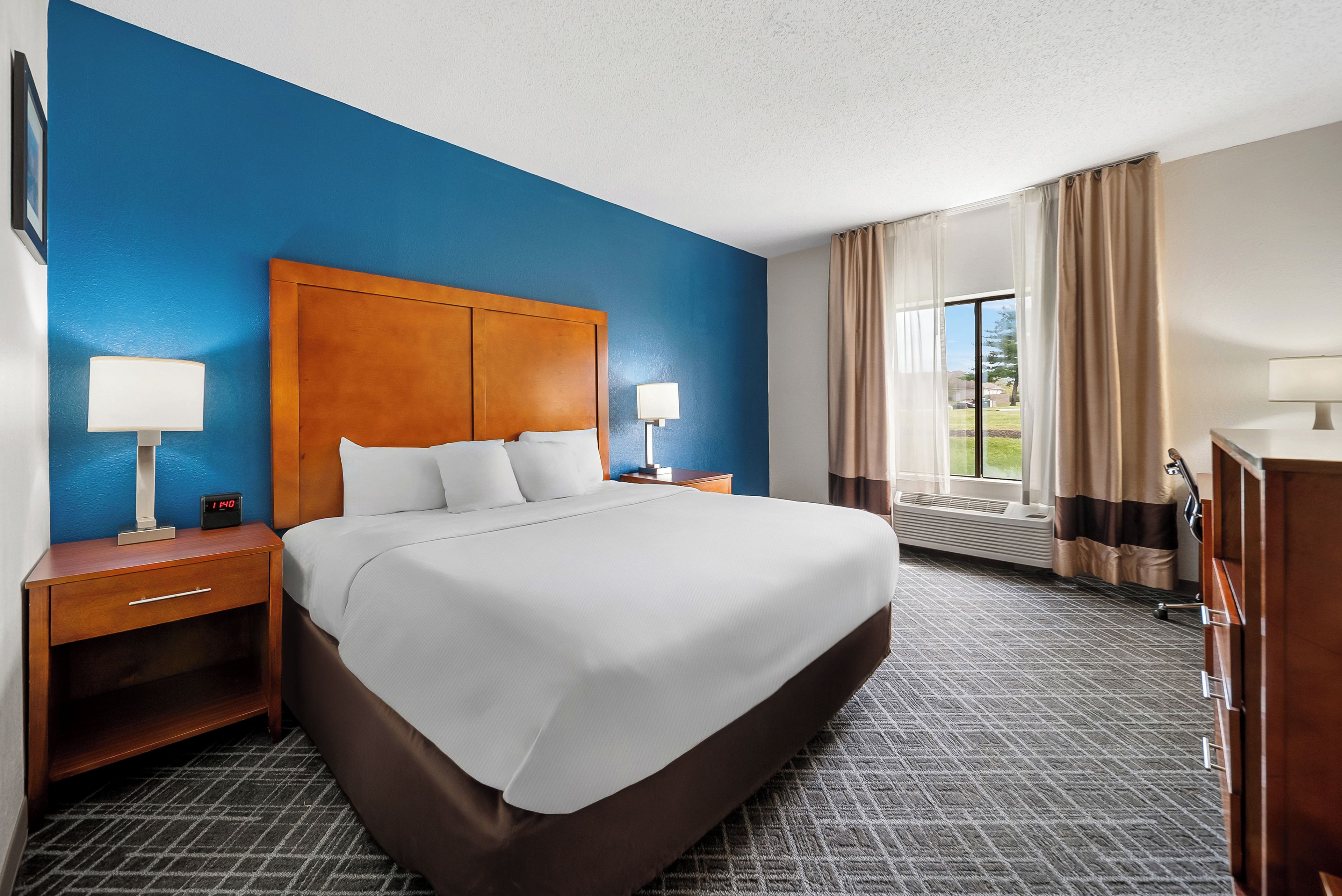 A hotel room with a large bed, two bedside tables with lamps, a nightstand, a dresser, and a window with curtains.