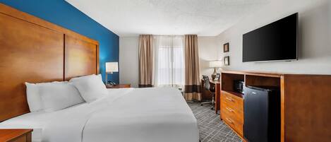 A hotel room with a large bed, a desk with a chair, a flat-screen TV, and a minibar.