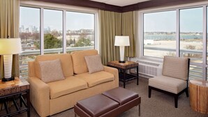 A hotel room with a beige sofa, a brown ottoman, a wicker chair, and a view of a cityscape and beach through large windows. - DoubleTree by Hilton Hotel Boston Bayside (Boston)
