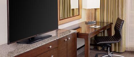 A hotel room with a flat-screen TV on a granite countertop, a wooden desk with a black office chair, a lamp, and a mirror.