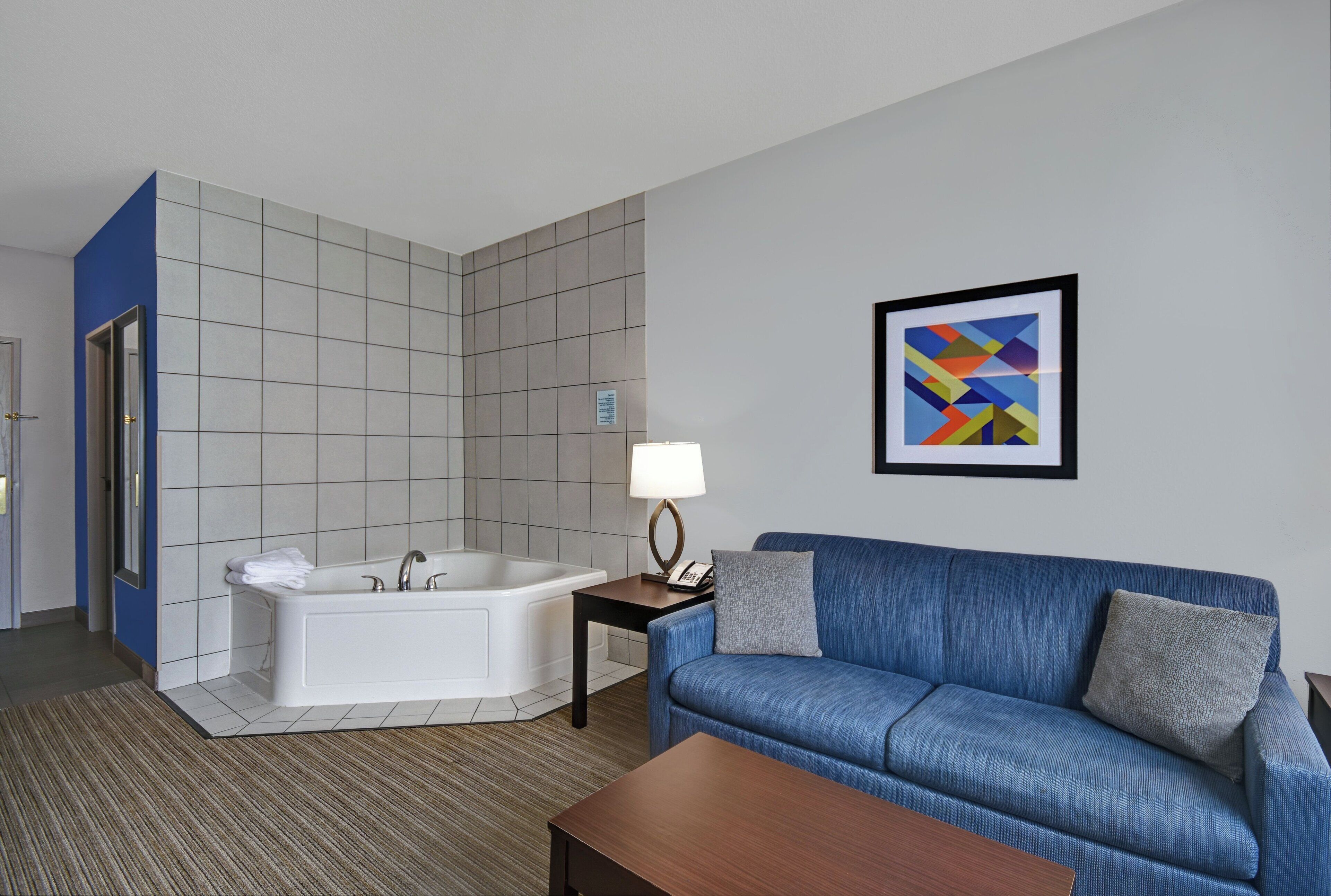 A hotel room with a bathtub, a blue sofa, a wooden coffee table, and a framed abstract painting on the wall.