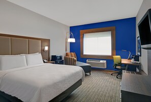 A hotel room with a large bed, a desk with a computer, a chair, a lamp, and a window with blinds.