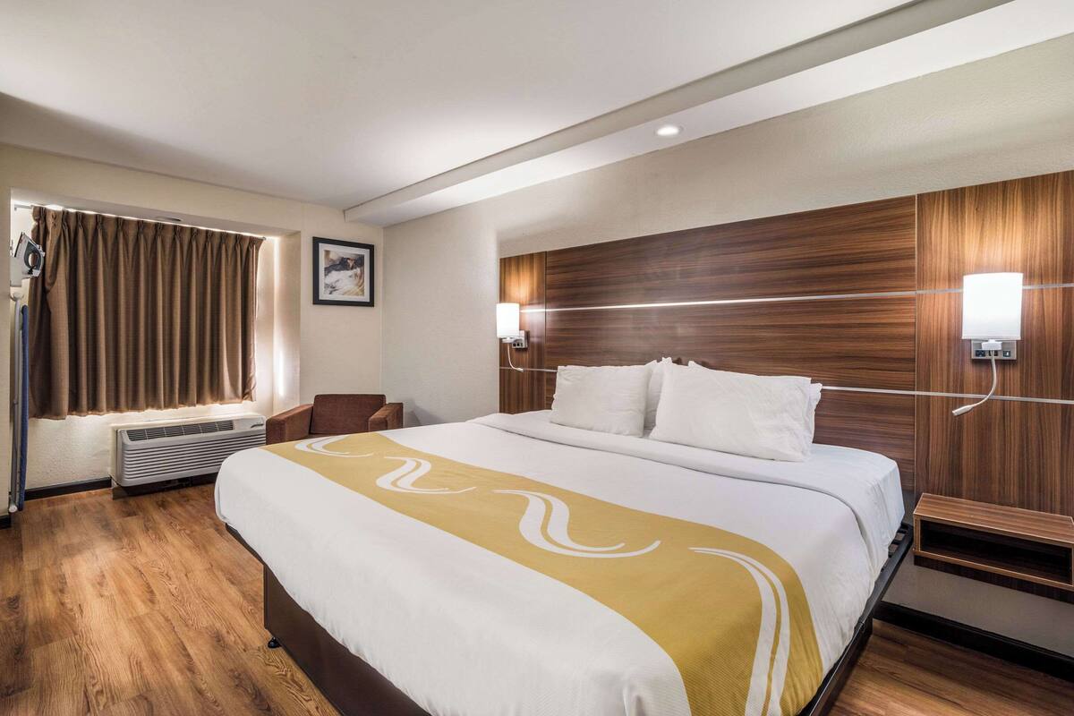 Una habitación de hotel con una cama grande, cabecera de madera, dos lámparas de noche, una ventana con cortinas y un aire acondicionado instalado en la pared.