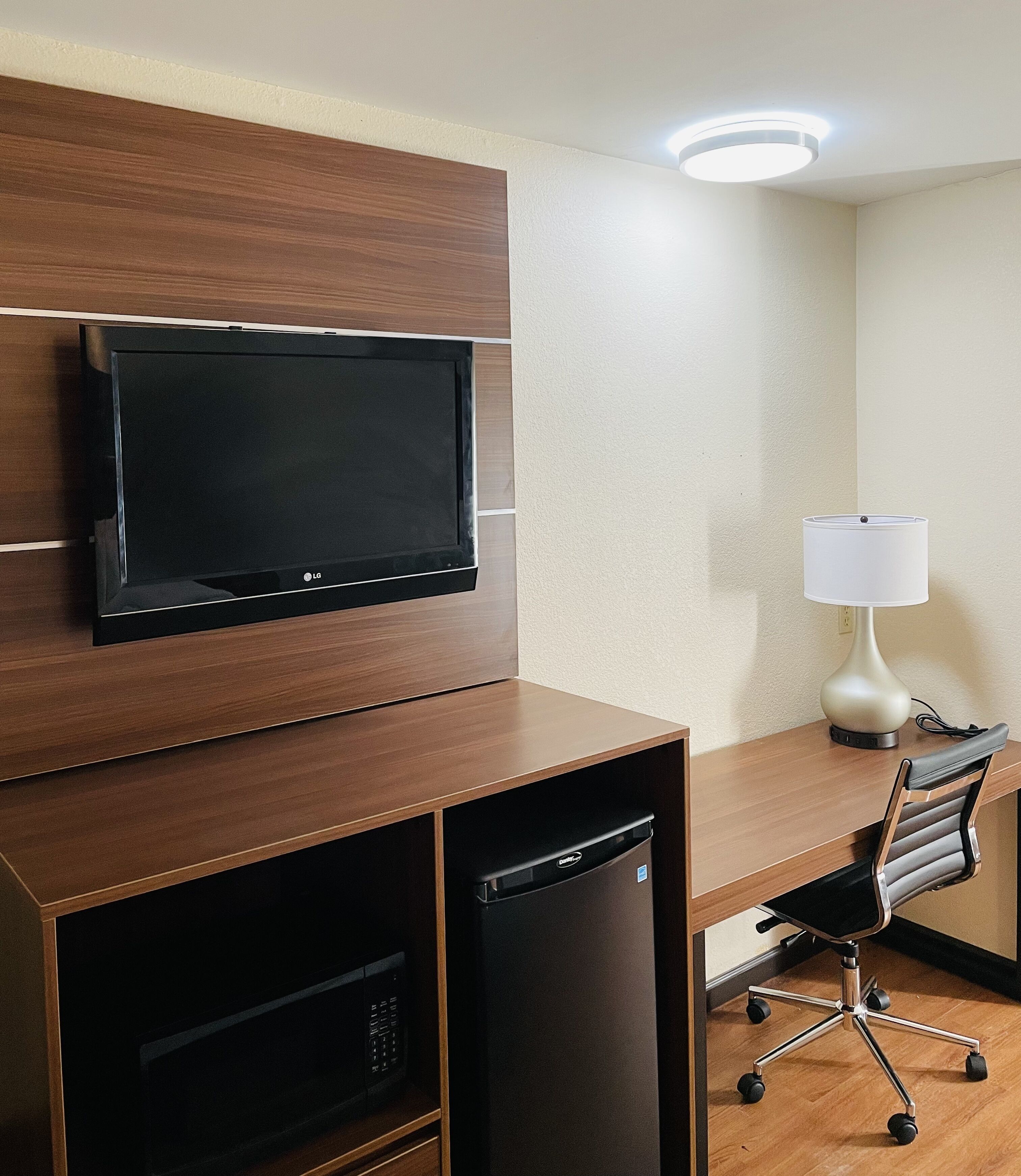 A hotel room with a flat-screen TV mounted on a wooden cabinet, a desk with a lamp, and a compact refrigerator.