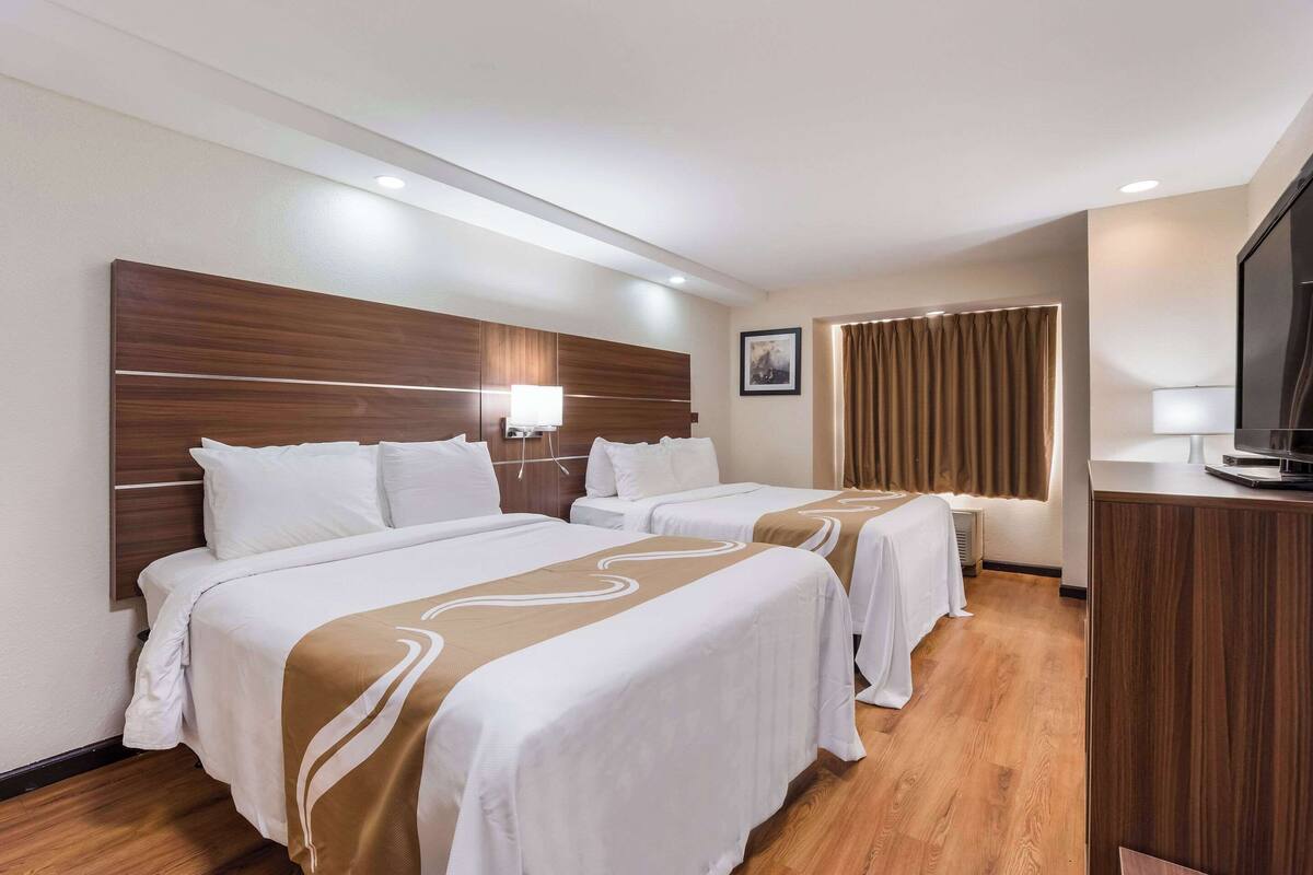 Una habitación de hotel con dos camas, un cabecero de madera, un televisor y una ventana con cortinas.