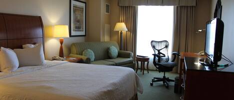 A hotel room with a large bed, a desk with a computer, a chair, a sofa, and a television.