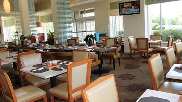 A restaurant with multiple tables and chairs, large windows, and a television mounted on the wall.