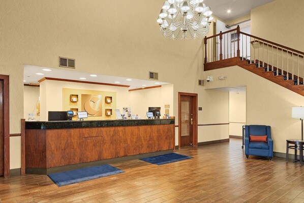 A hotel reception area with a wooden reception desk, a chandelier, a staircase, and a blue armchair. - Comfort Suites Milwaukee Airport (Oak Creek)