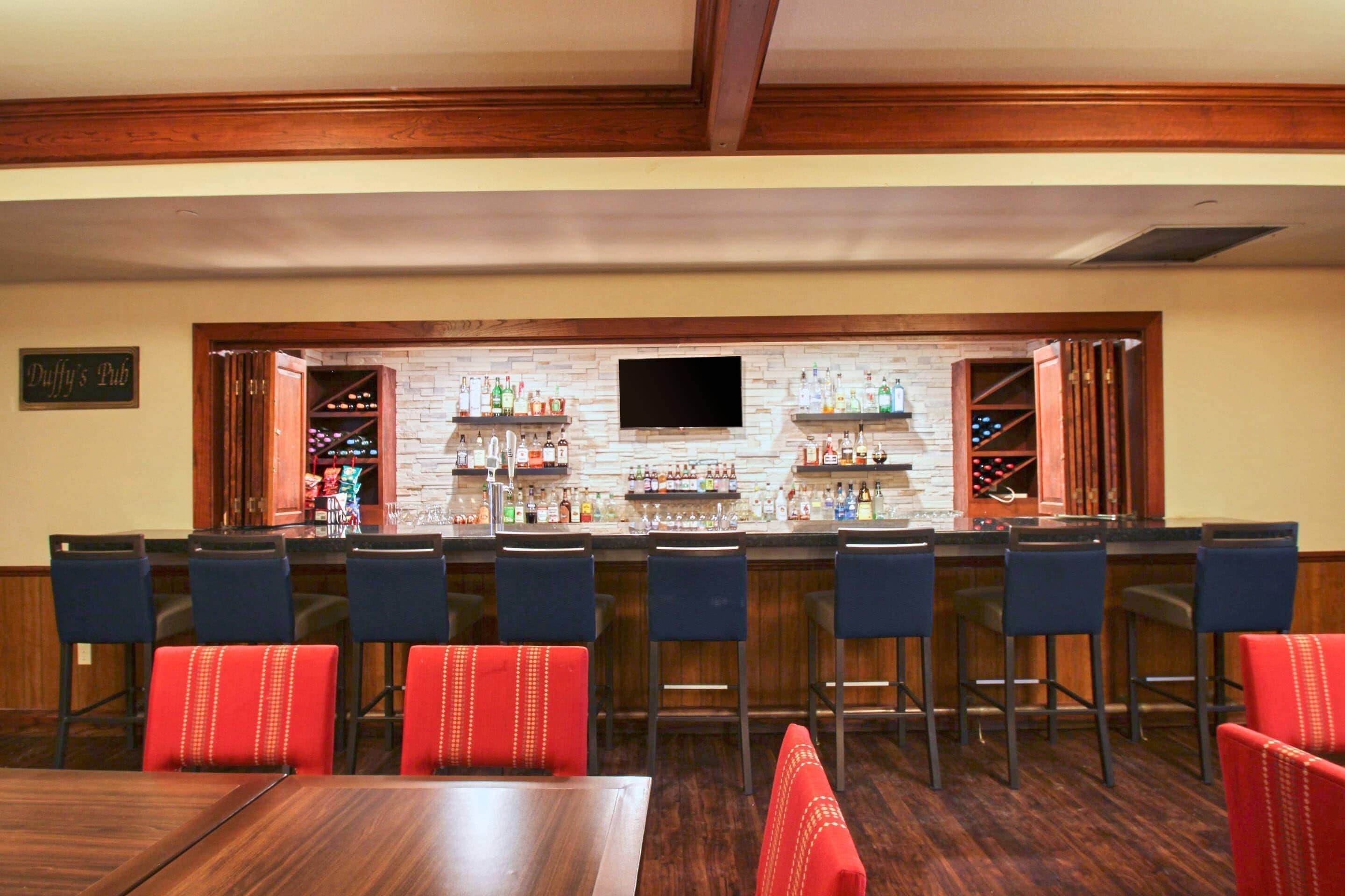 Zona de bar con pared de ladrillo, televisor de pantalla plana y varias botellas. Hay taburetes de bar y una mesa de comedor con sillas.