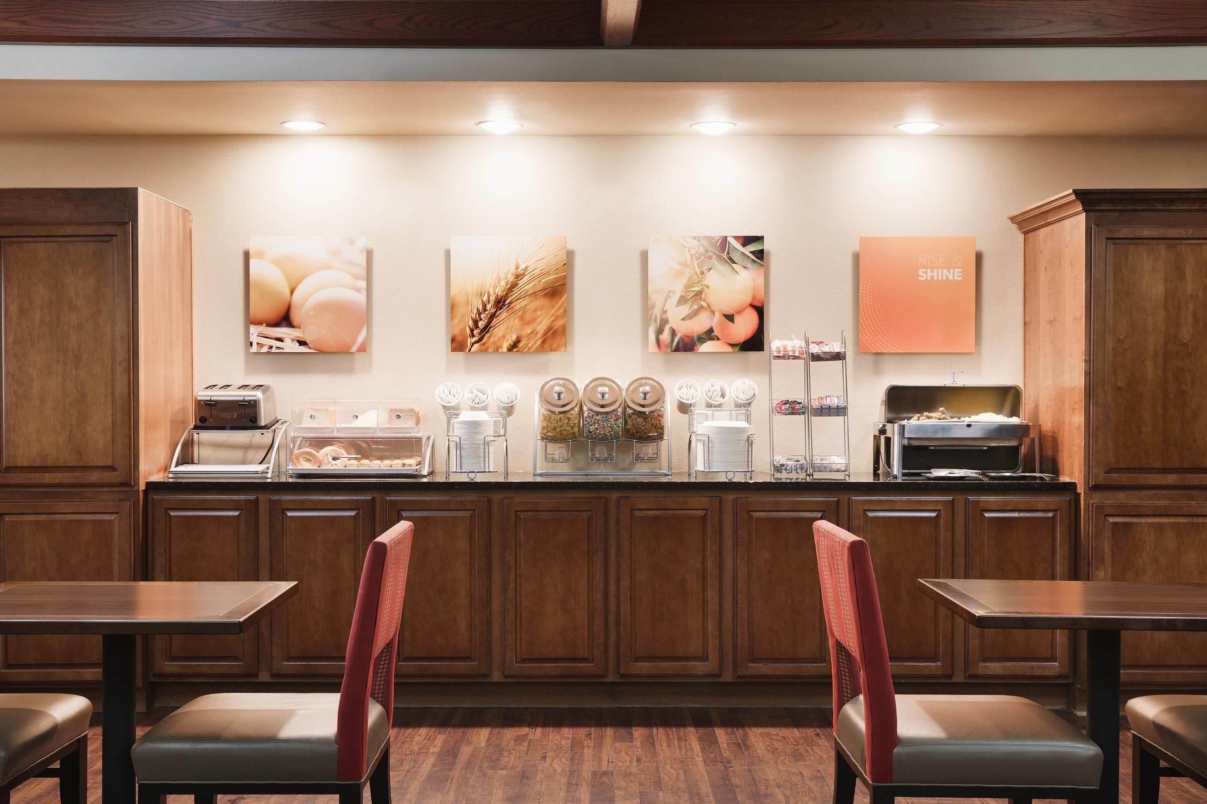 A breakfast bar with a coffee machine, a toaster, and a fruit display. There are wooden cabinets and a sign that says 'Rise & Shine'.