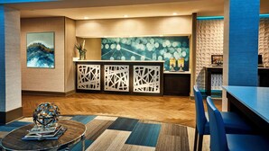 A modern hotel lobby with a wooden herringbone floor, a reception desk with patterned partitions, a large abstract painting, and a round table with magazines. - Best Western Plus Lewisville Flower Mound (Lewisville)