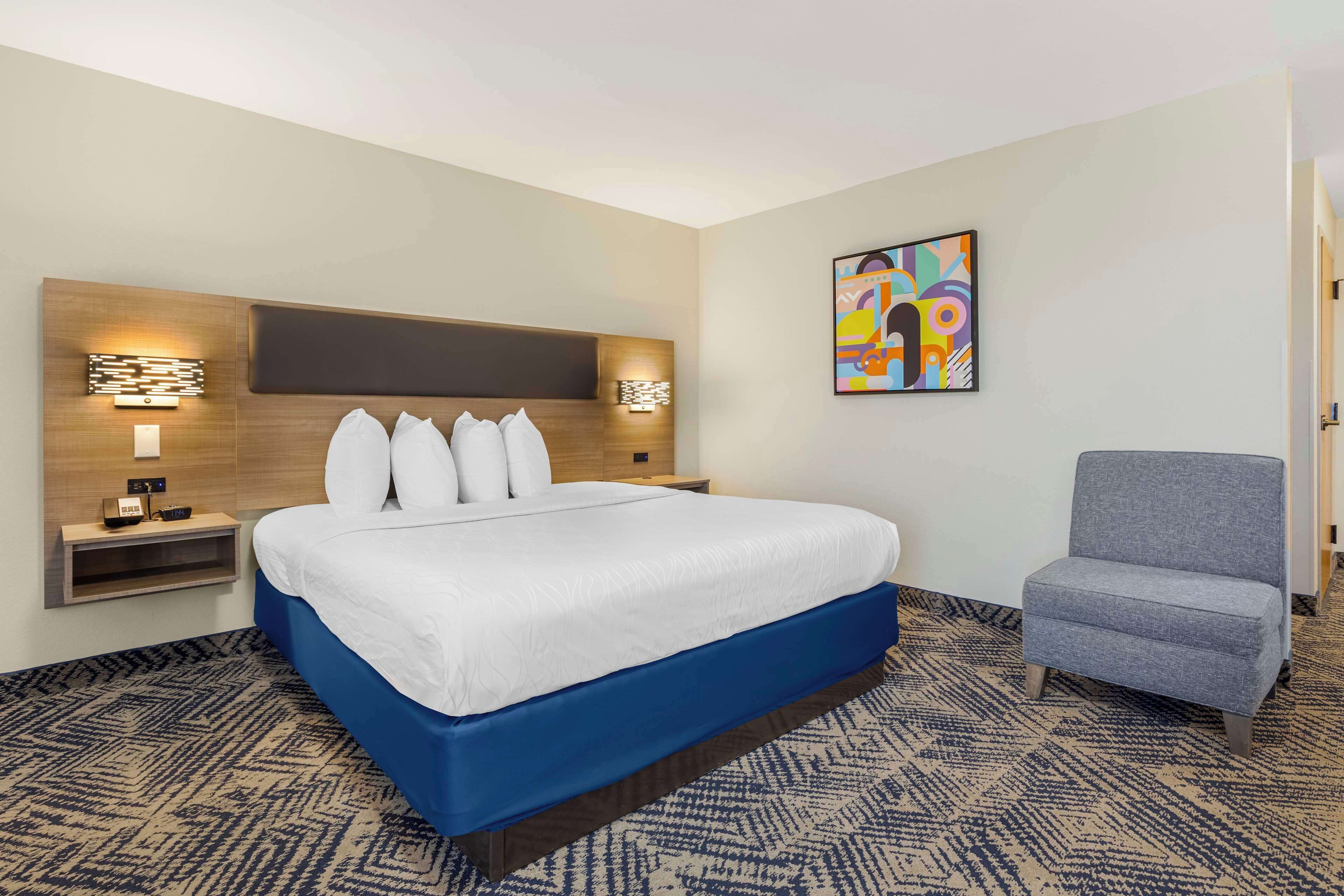 Ein Hotelzimmer mit einem großen Bett, einem Nachttisch mit Lampe, einem Sessel und einem farbenfrohen, abstrakten Gemälde an der Wand.