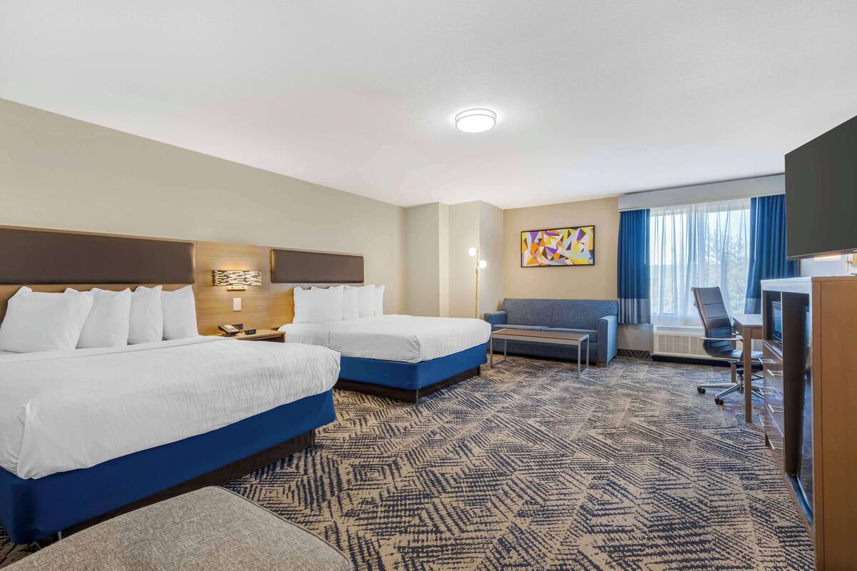 A hotel room with two beds, a desk, a chair, a sofa, and a large window with curtains.