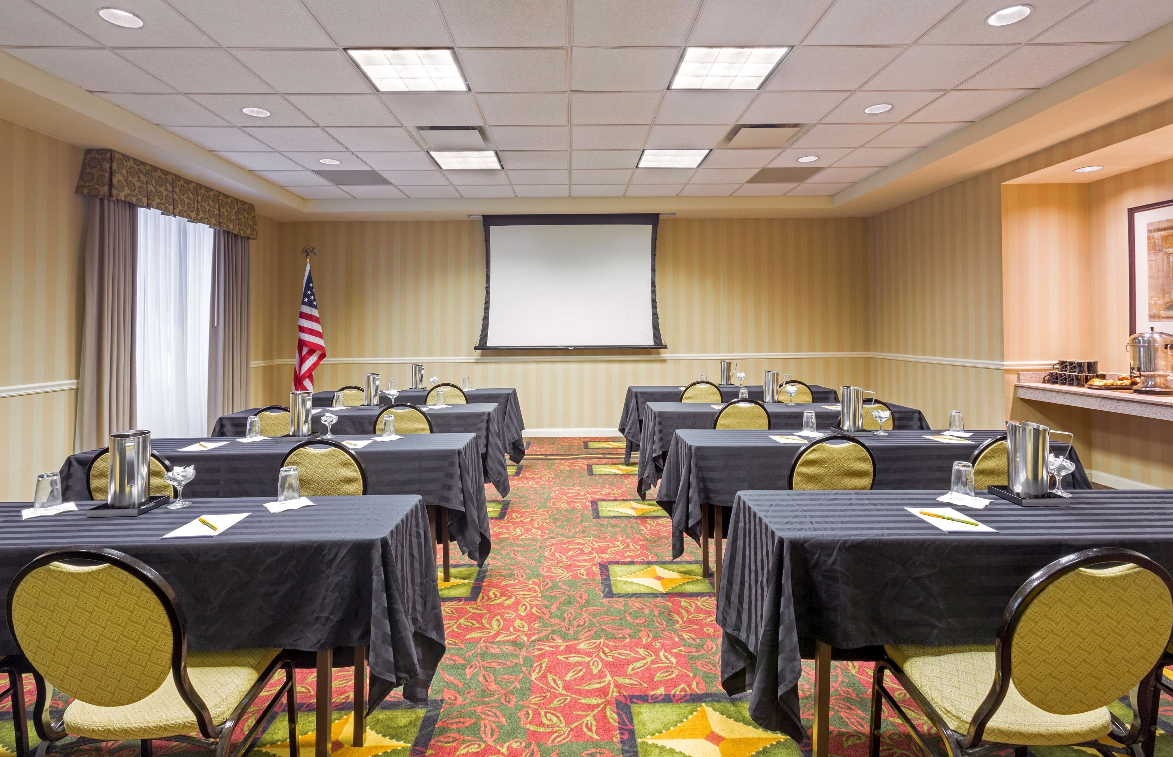 A conference room with multiple tables, chairs, a projector screen, and an American flag.