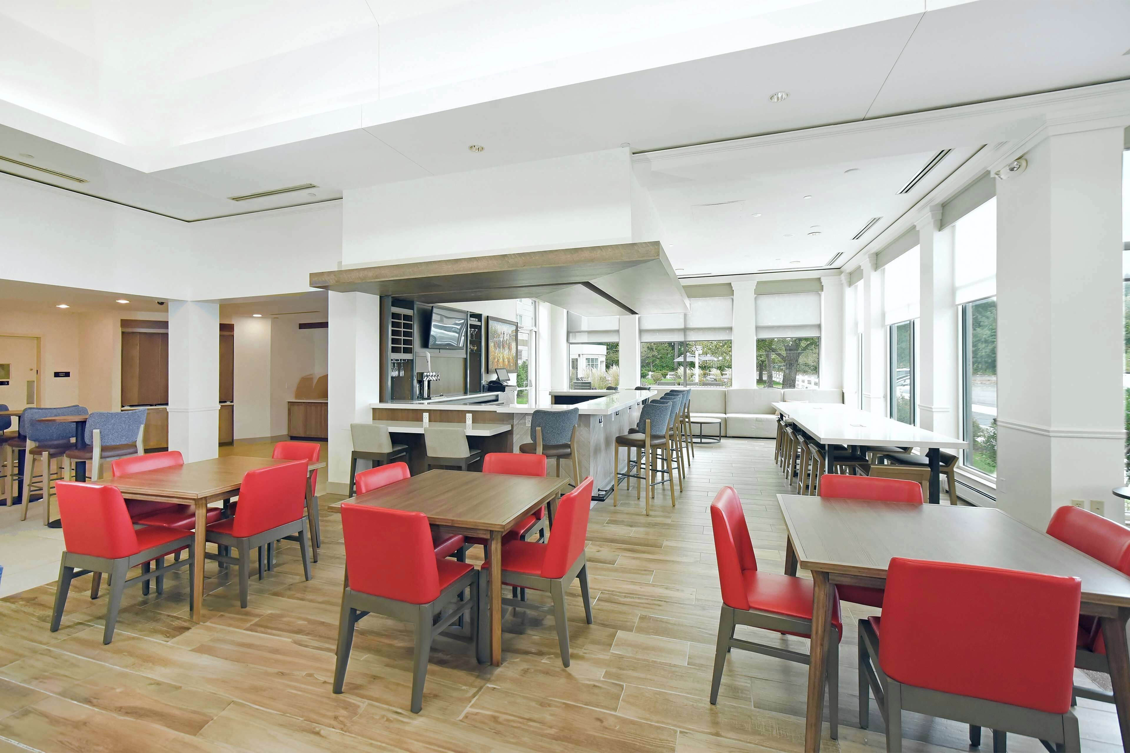 Un vaste espace intérieur aménagé en salle à manger, avec de nombreuses tables et chaises, un coin cuisine et de grandes fenêtres laissant entrer la lumière naturelle.