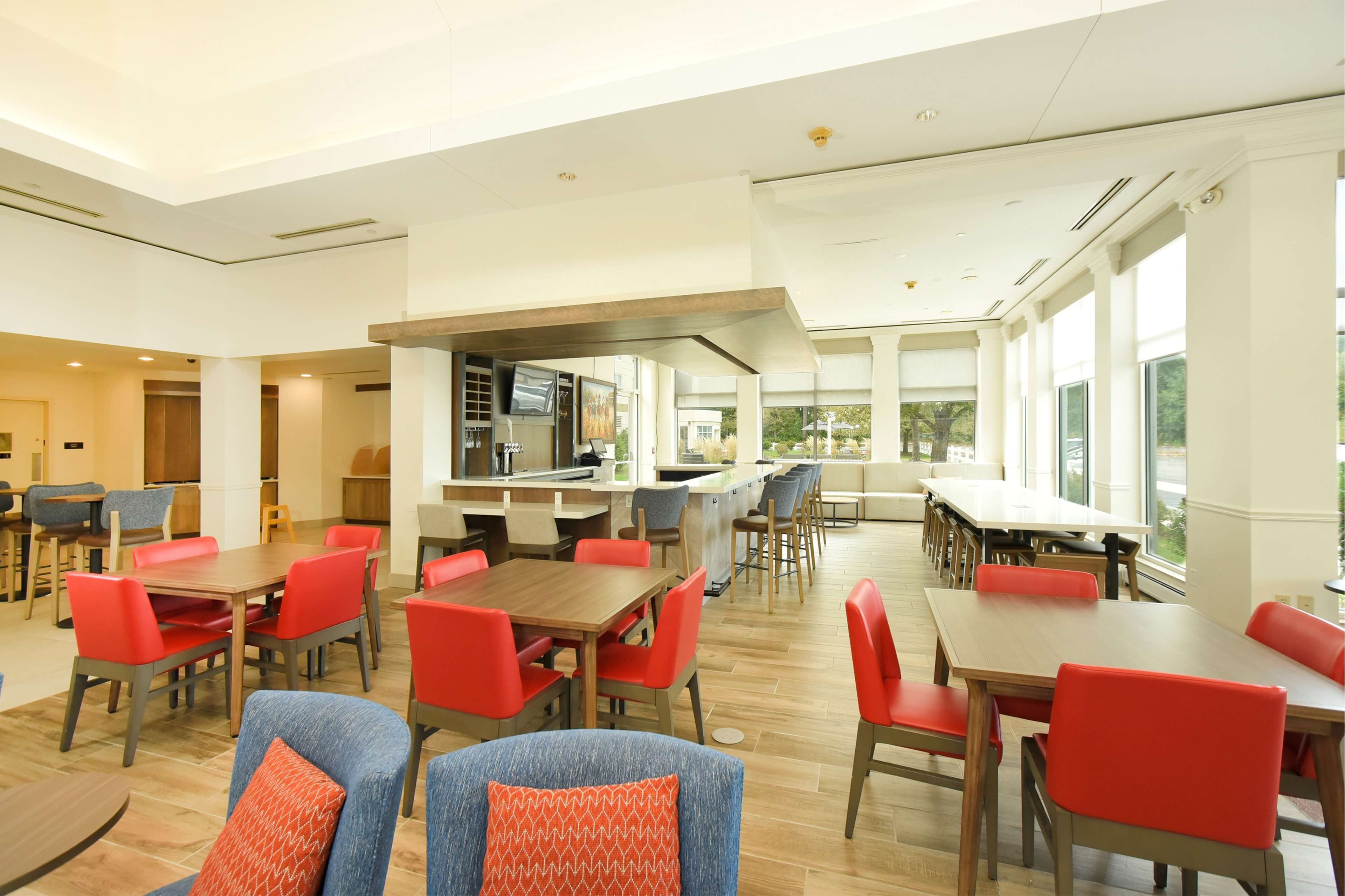 A spacious dining area with multiple tables and chairs, a kitchen area, and large windows providing natural light.