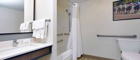 A bathroom with a sink, mirror, shower, toilet, and a mounted picture of a horse race.