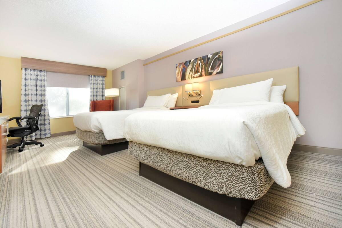 Habitación de hotel con dos camas, un escritorio con silla, una ventana con cortinas y una obra de arte en la pared.