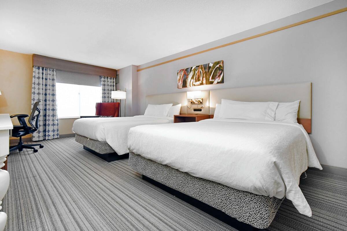 Una habitación de hotel con dos camas, un escritorio, una silla y un ventanal con cortinas.