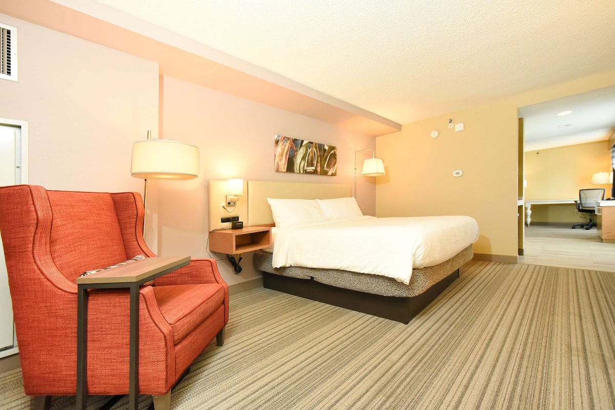 Una habitación de hotel con una cama, un sillón rojo, una mesita, una mesita de noche y una lámpara.