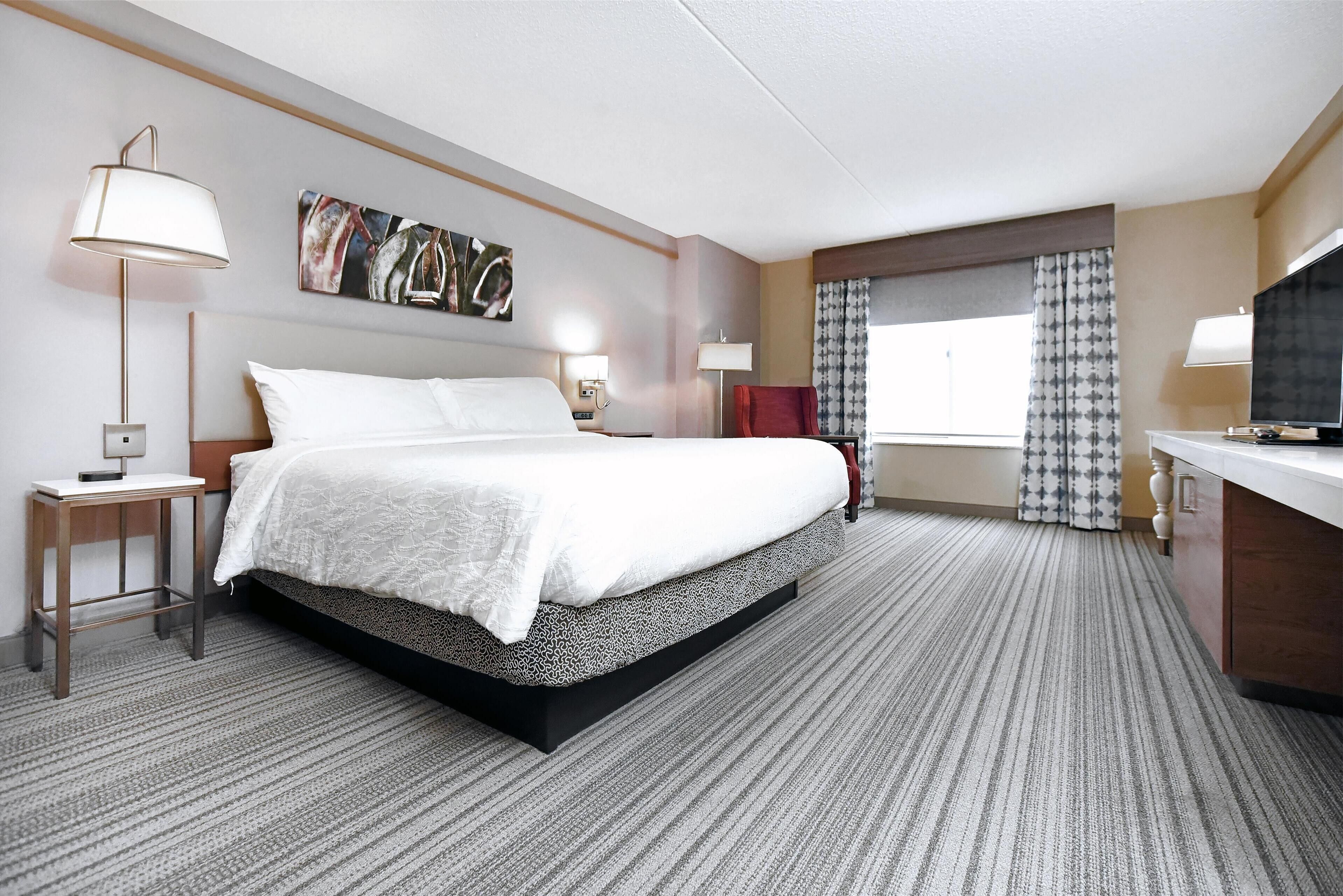 A hotel room with a large bed, two bedside tables with lamps, a flat-screen TV, a red chair, and a window with patterned curtains.