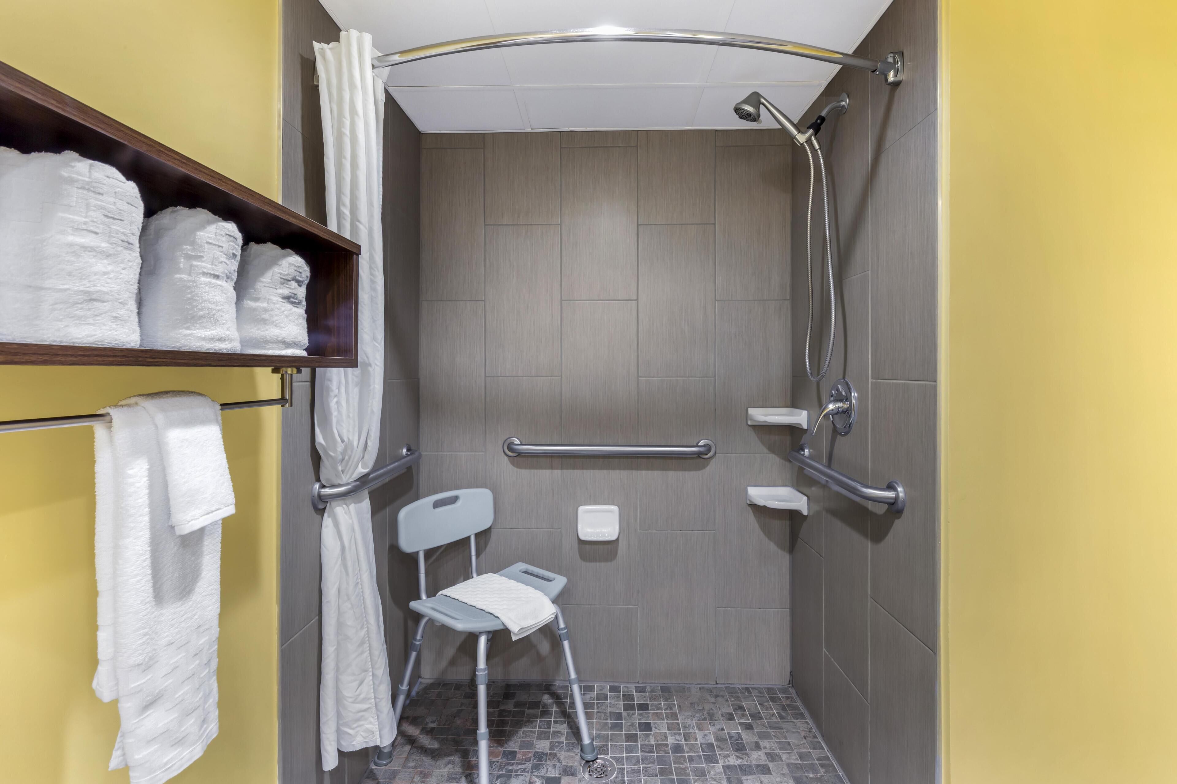 A shower area with a grab bar, a shelf with towels, and a small chair.
