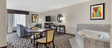A hotel room with a dining area, a TV, a desk, a chair, a sofa, a large mirror, a lamp, and a bathtub.