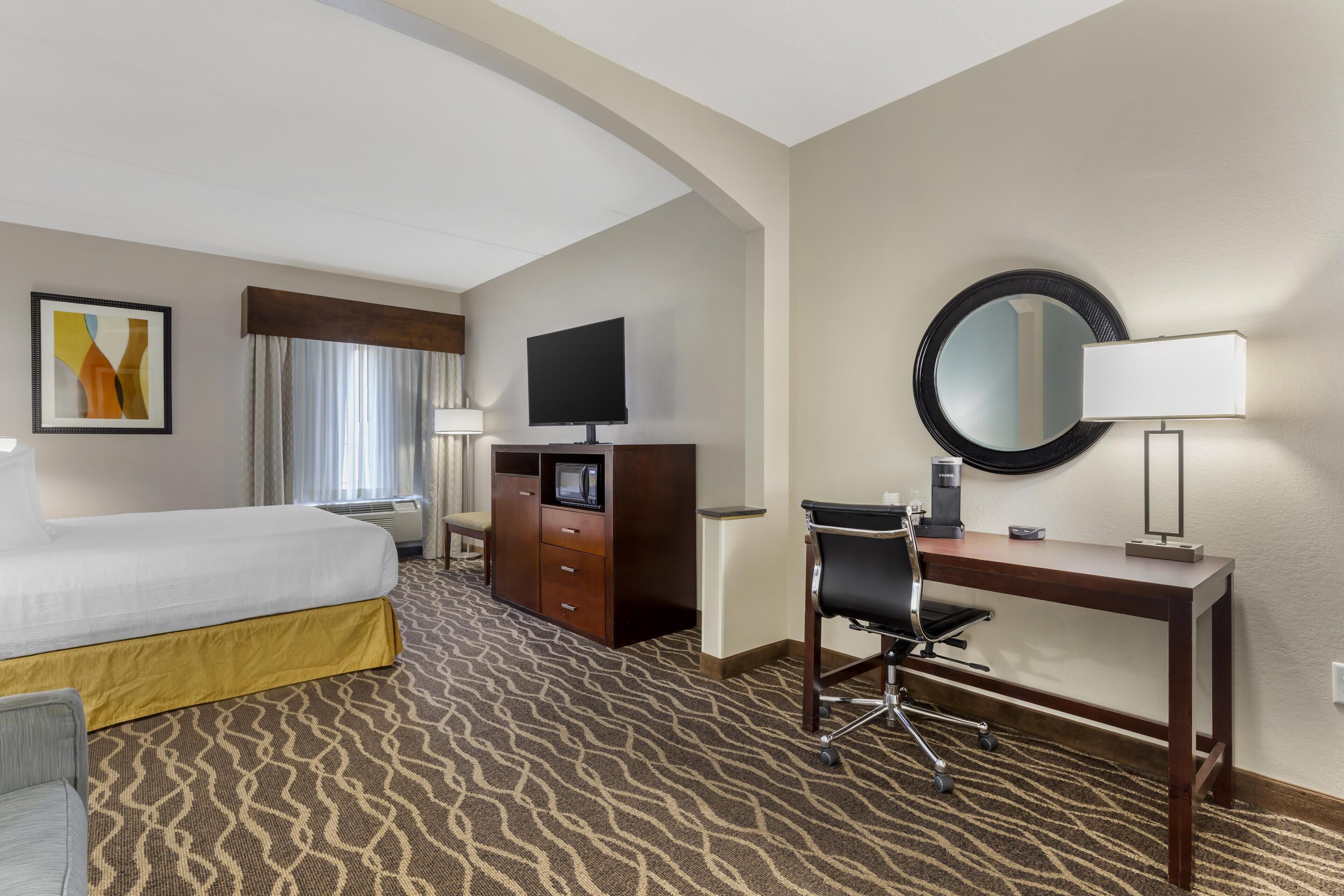 Habitación de hotel con cama, escritorio, silla, televisor, cómoda y un espejo grande.