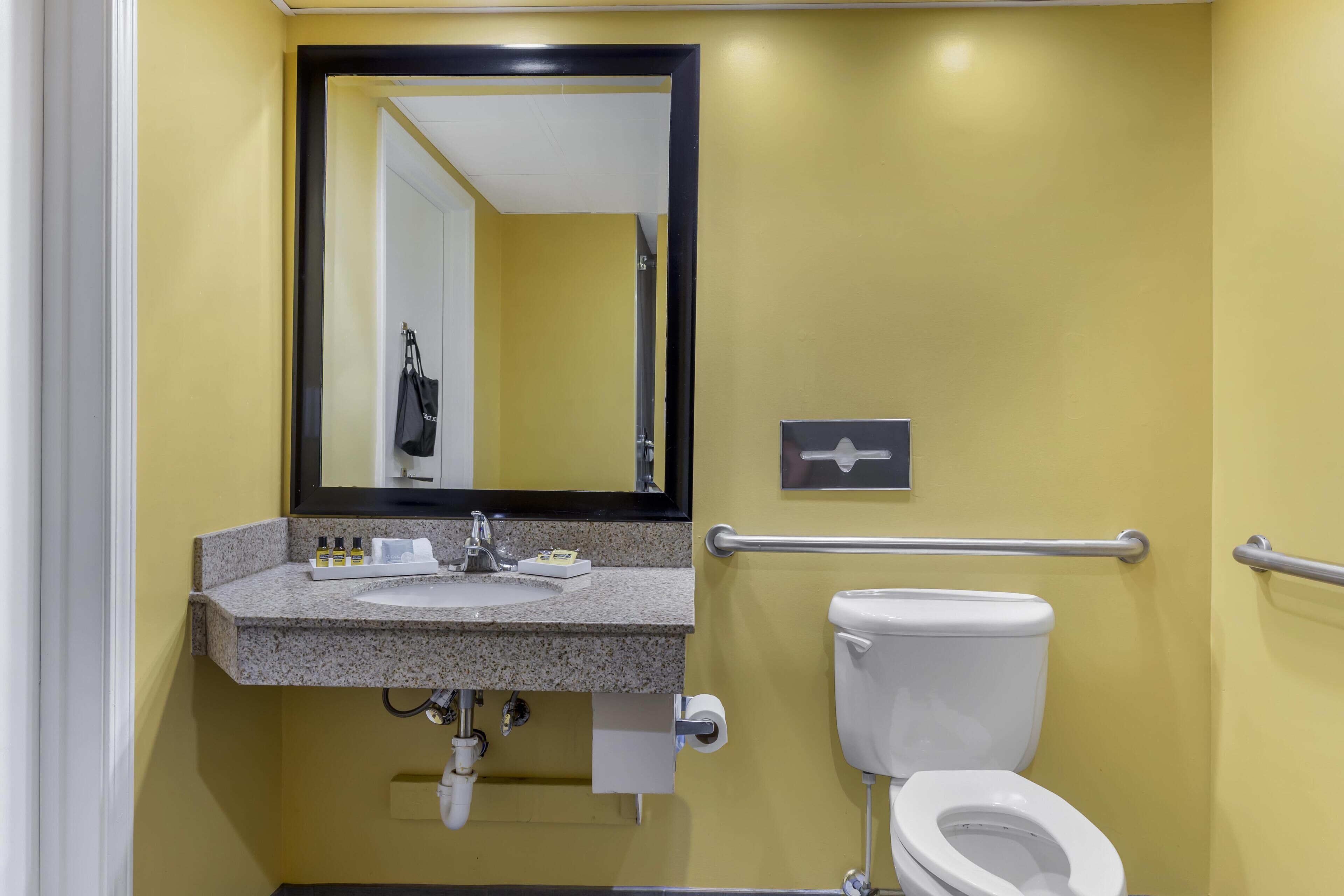 A bathroom with a yellow wall, a granite countertop sink, a white toilet, a mirror, and a handrail.