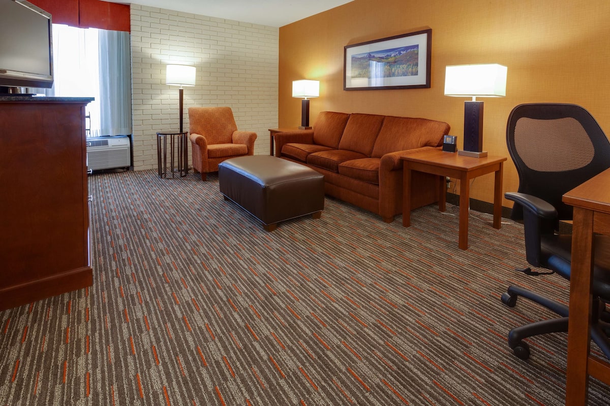 A hotel room with a brown sofa, a wooden table, a black office chair, a small ottoman, and two floor lamps.