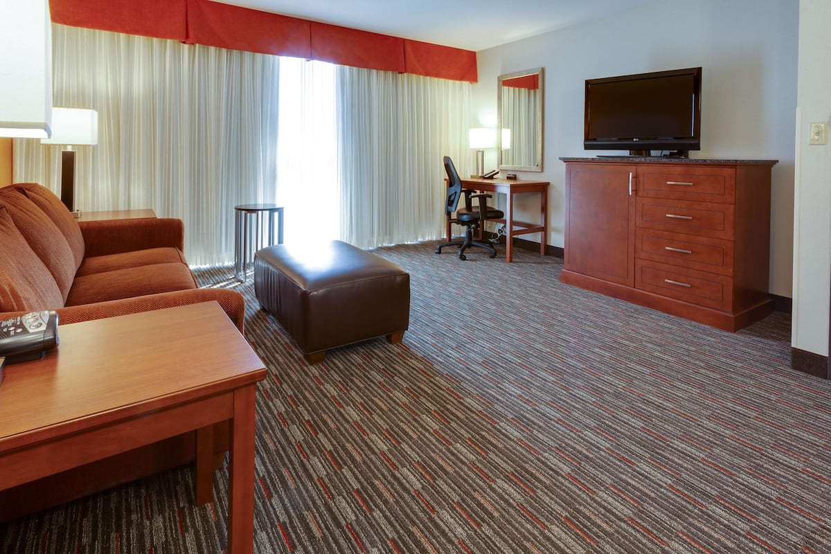 A hotel room with a sofa, ottoman, desk, television, and a large window with curtains.