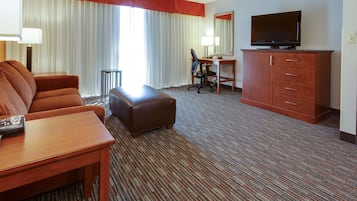 A hotel room with a sofa, ottoman, desk, television, and a large window with curtains.