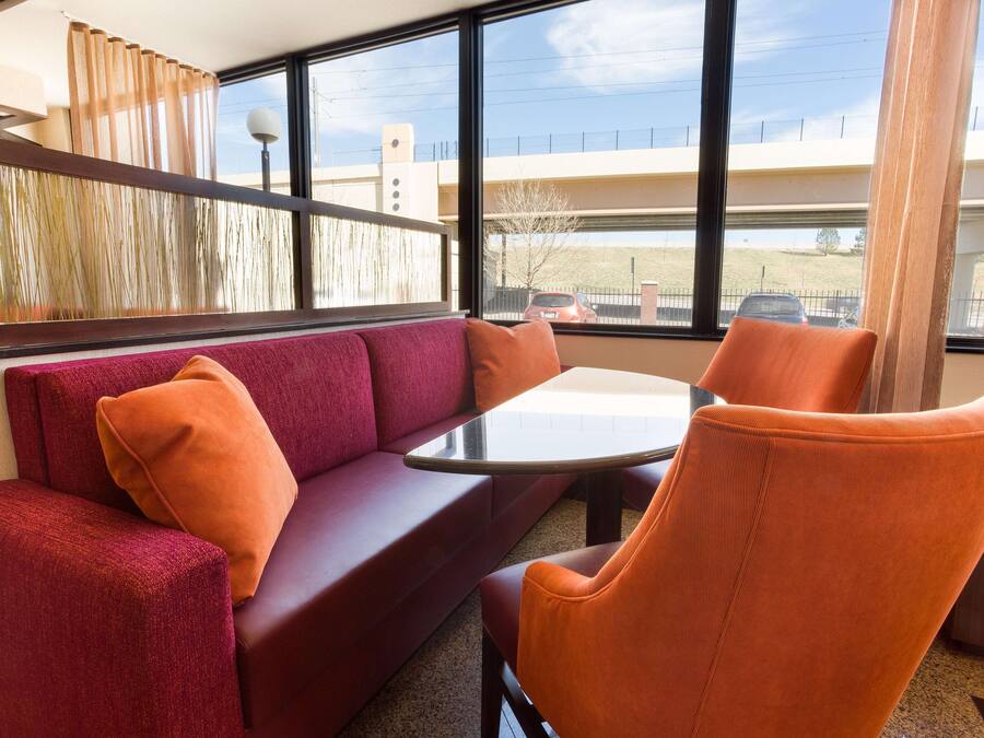 Une pièce moderne avec un canapé rouge, des fauteuils orange, une table en verre et de grandes fenêtres donnant sur un immeuble et un parking.