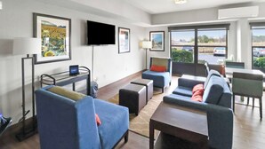 A modern hotel room with a sofa set, a flat-screen TV, artwork on the walls, and large windows offering a view of the parking area.