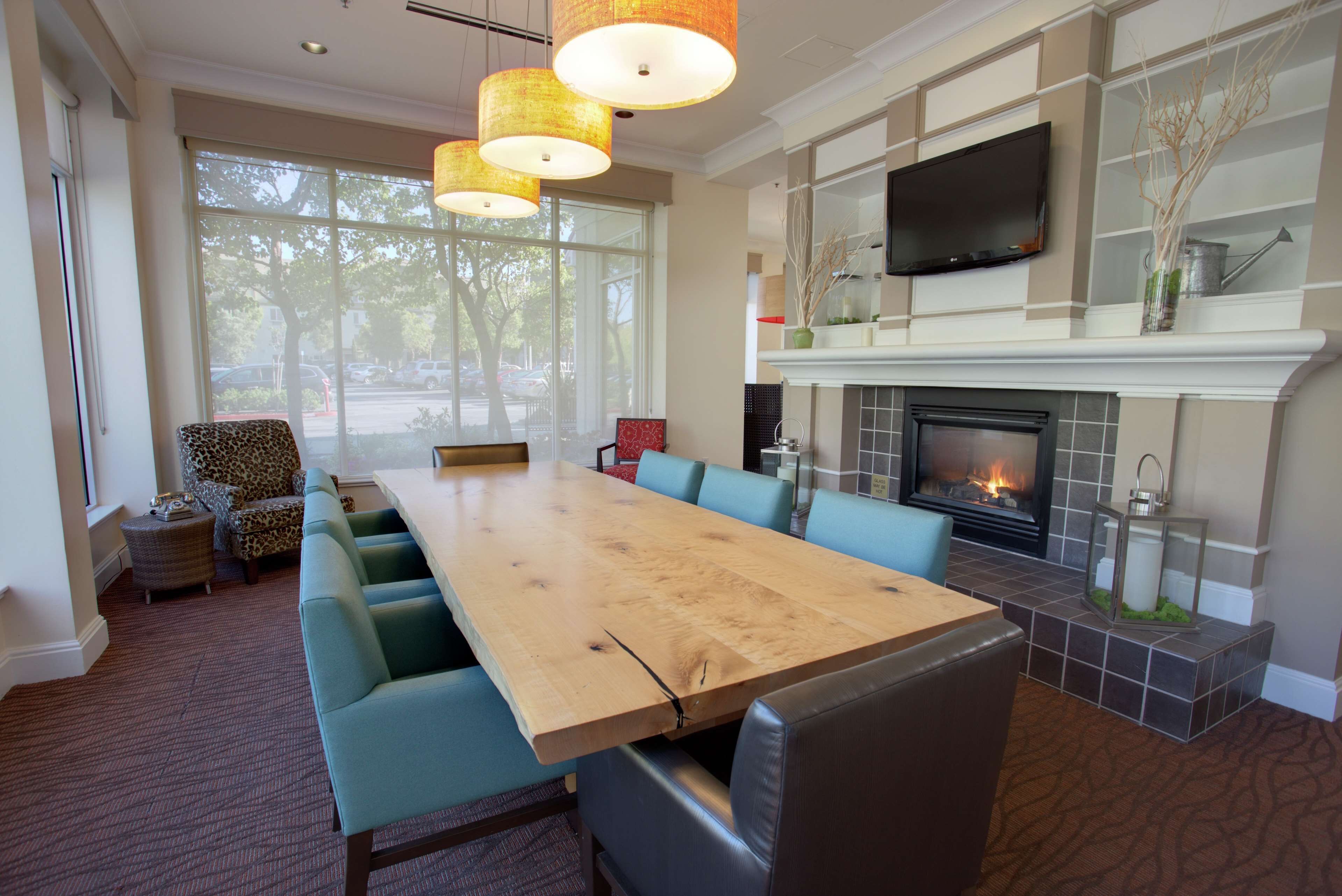 A spacious dining area with a long wooden table and blue upholstered chairs, a fireplace, and a television mounted above it.