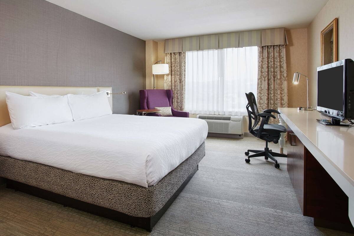 Ein Hotelzimmer mit einem großen Bett, einem Schreibtisch mit Computer, einem Sessel und einem Fernseher.