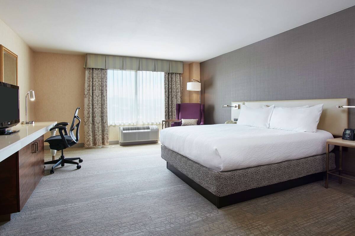 Ein Hotelzimmer mit einem großen Bett, einem Schreibtisch mit Computer, einem Sessel, einem Fernseher und einem Fenster mit Vorhängen.