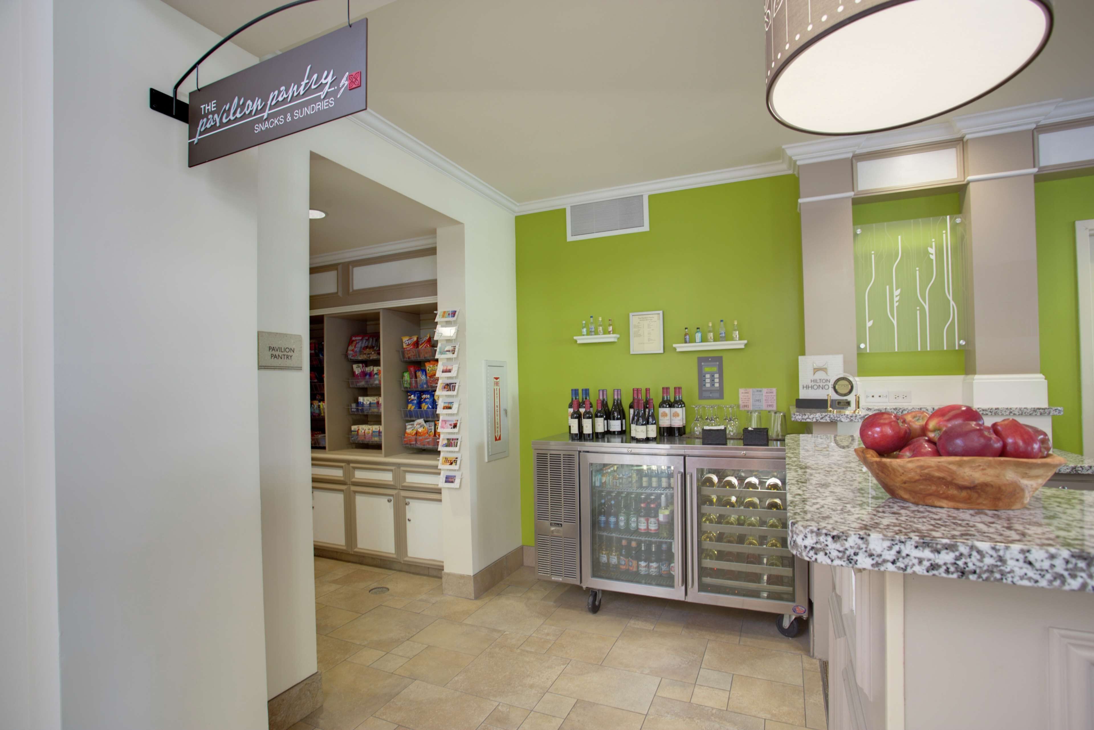 A hotel pantry with snacks and sundries, a refrigerated display case, and a fruit bowl on the counter.