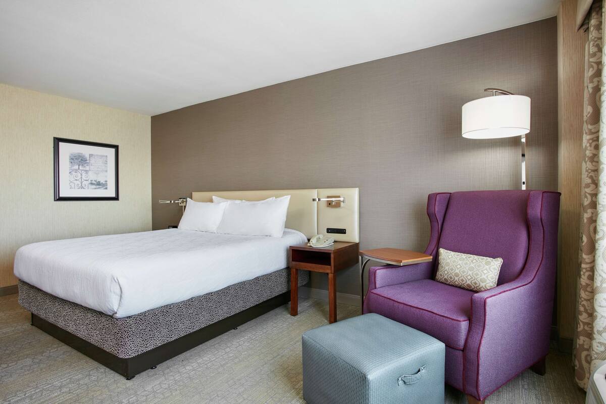Ein Hotelzimmer mit einem großen Bett, einem kleinen Tisch, einem violetten Sessel und einer Lampe.