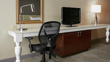 Una habitación de hotel con un escritorio blanco, una silla de oficina negra, un televisor, una lámpara y un espejo.