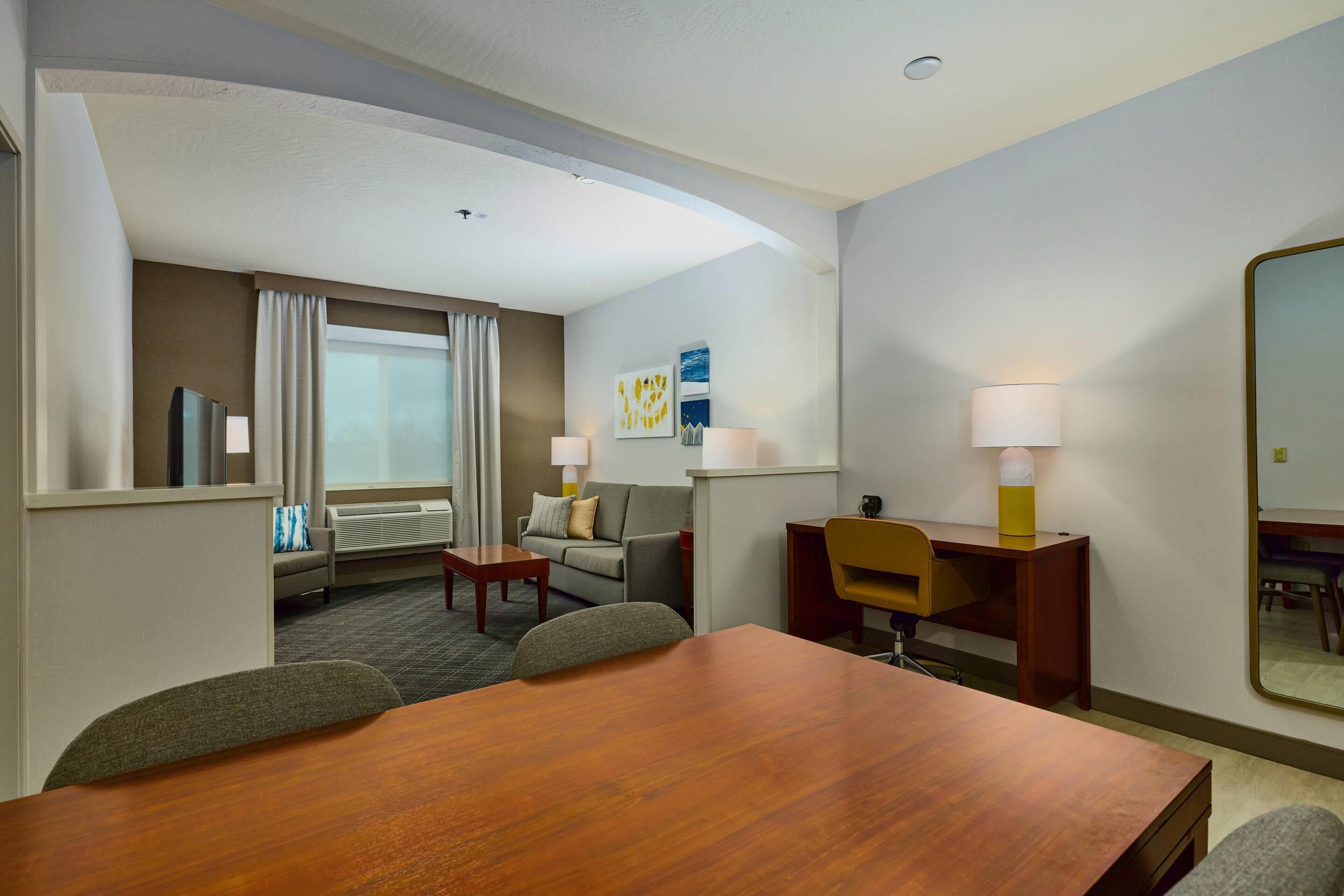 A hotel room with a wooden dining table, a sofa, a desk, and a large mirror.