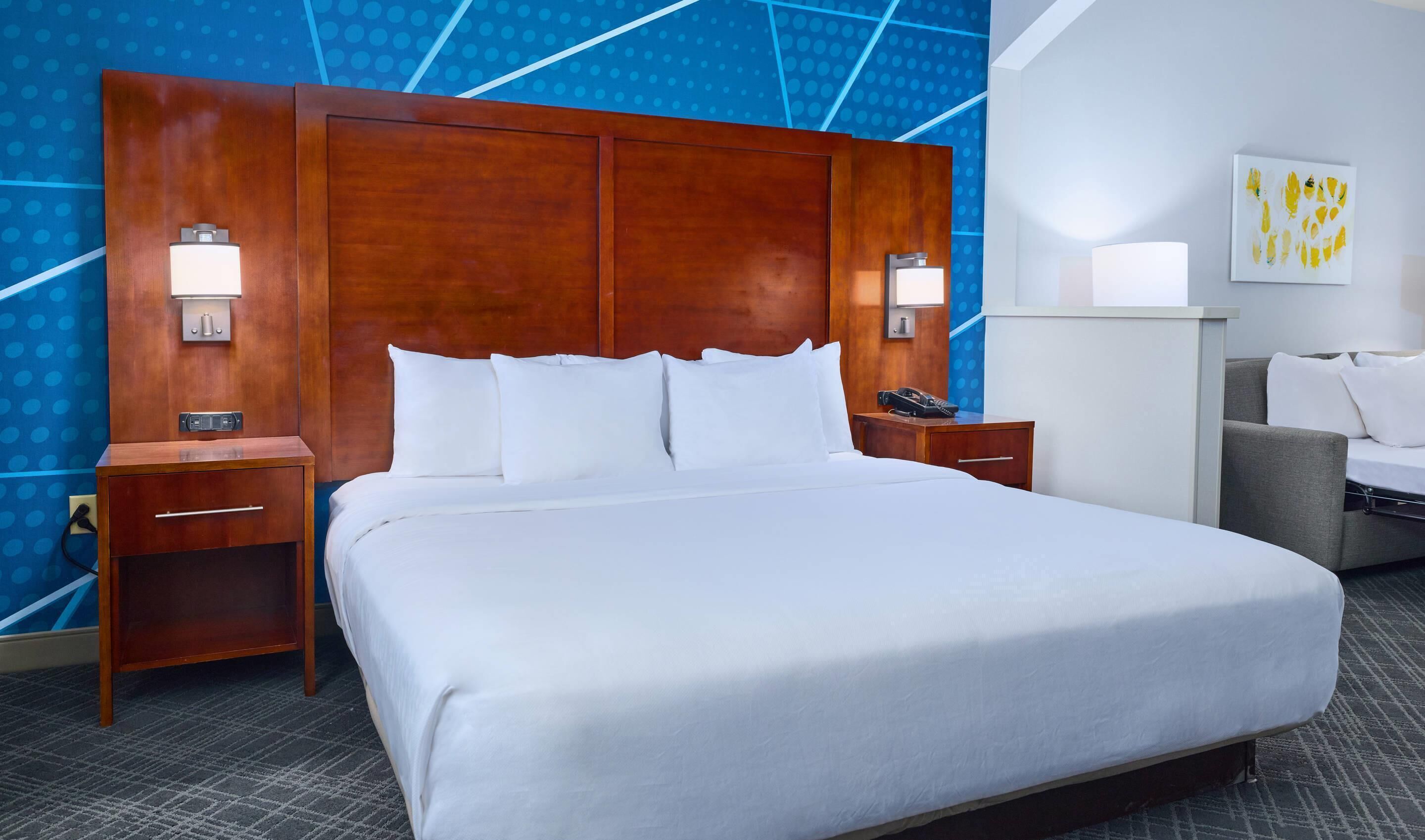 A hotel room with a large bed, two bedside tables, a desk with a chair, and a wall with a geometric pattern.