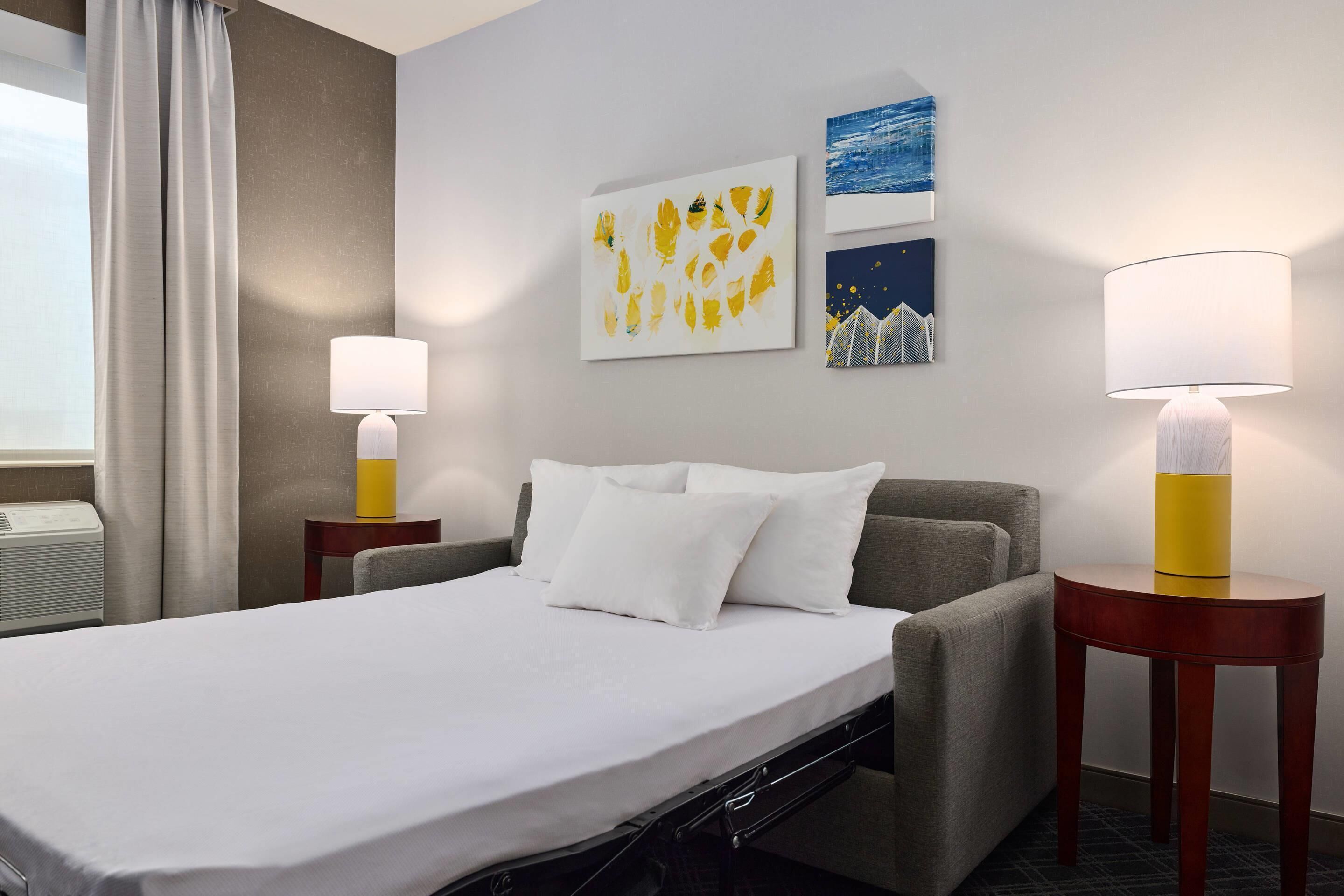 A hotel room with a bed, two bedside tables, a window with curtains, and artwork on the wall.