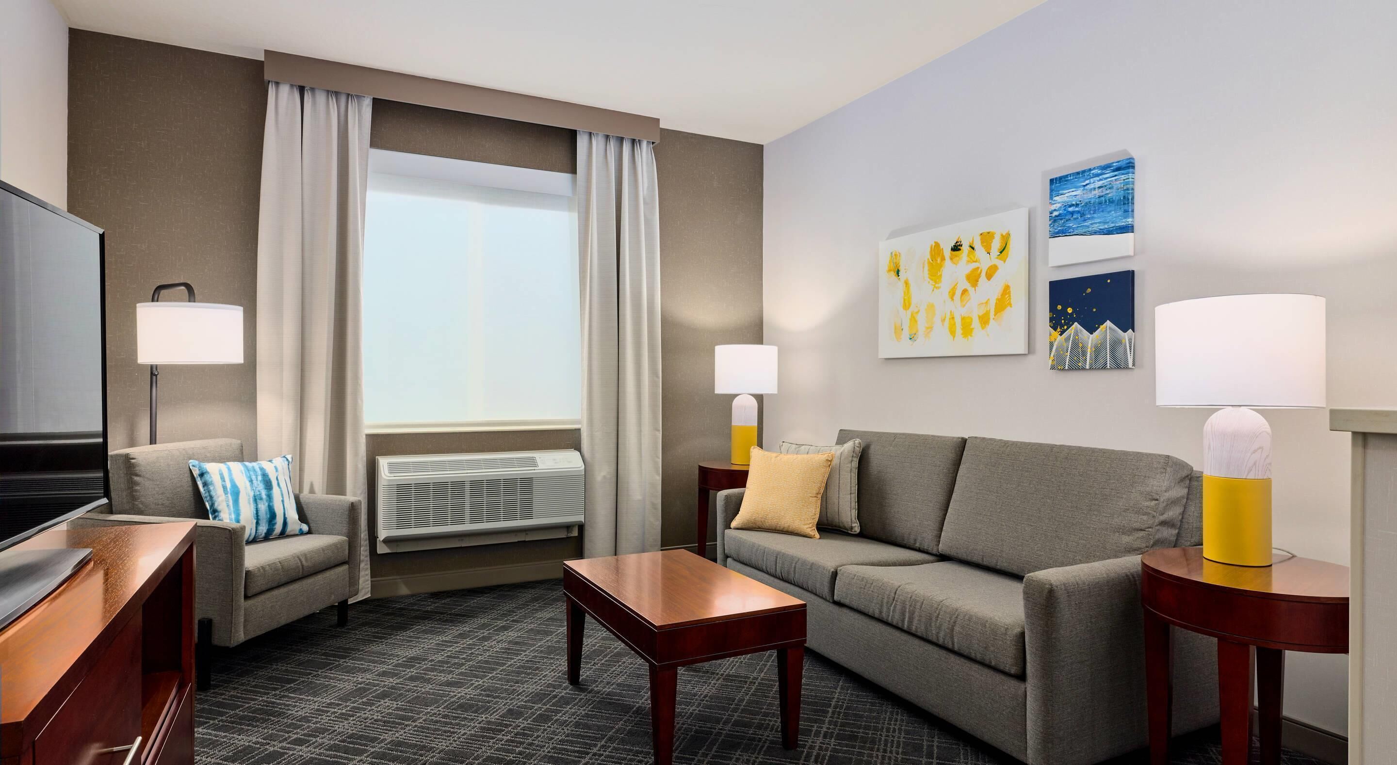 Ein Hotelzimmer mit einer Couch, zwei Sesseln, einem Fernseher, einem Flachbildfernseher, einem Couchtisch, zwei Lampen, einem Fenster mit Vorhängen und Kunstwerken an der Wand.