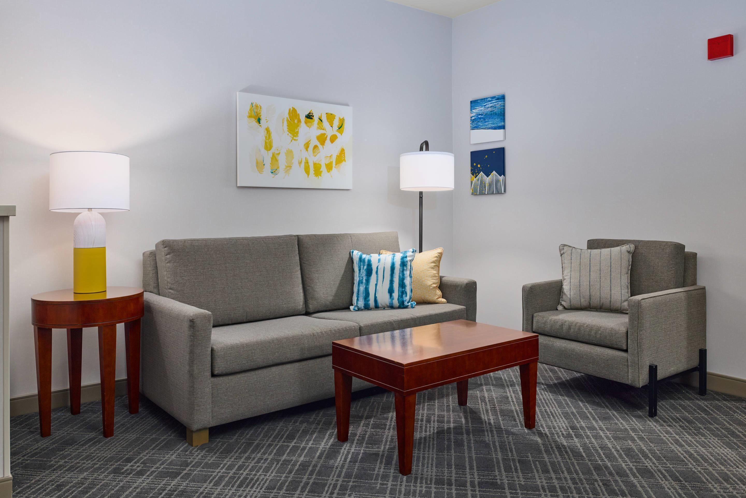 A hotel room with a grey sofa, a wooden coffee table, a side table with a lamp, and a chair. There are wall art pieces above the sofa.