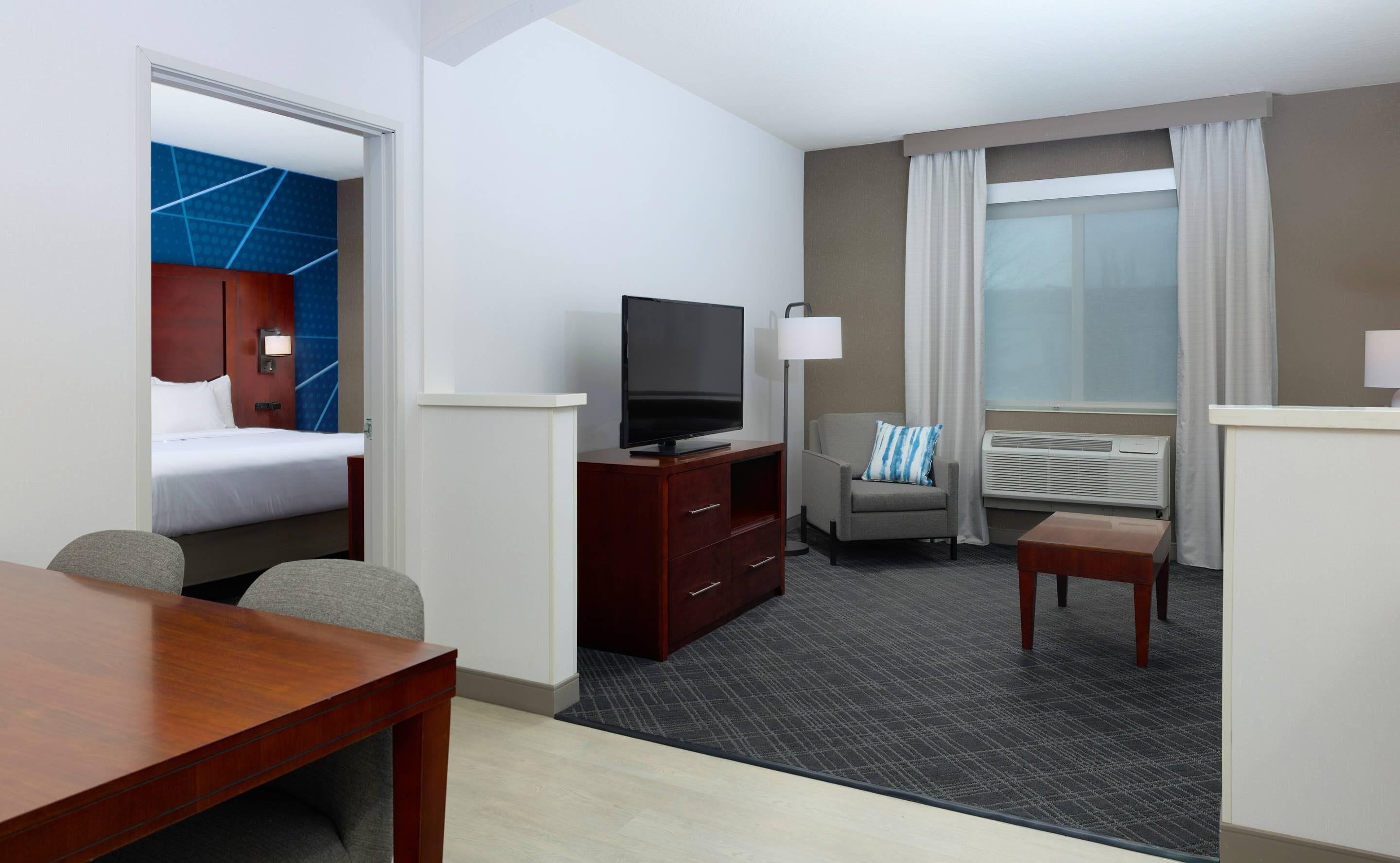 A hotel room with a bed, a TV, a desk, a chair, a sofa, and a window with curtains.