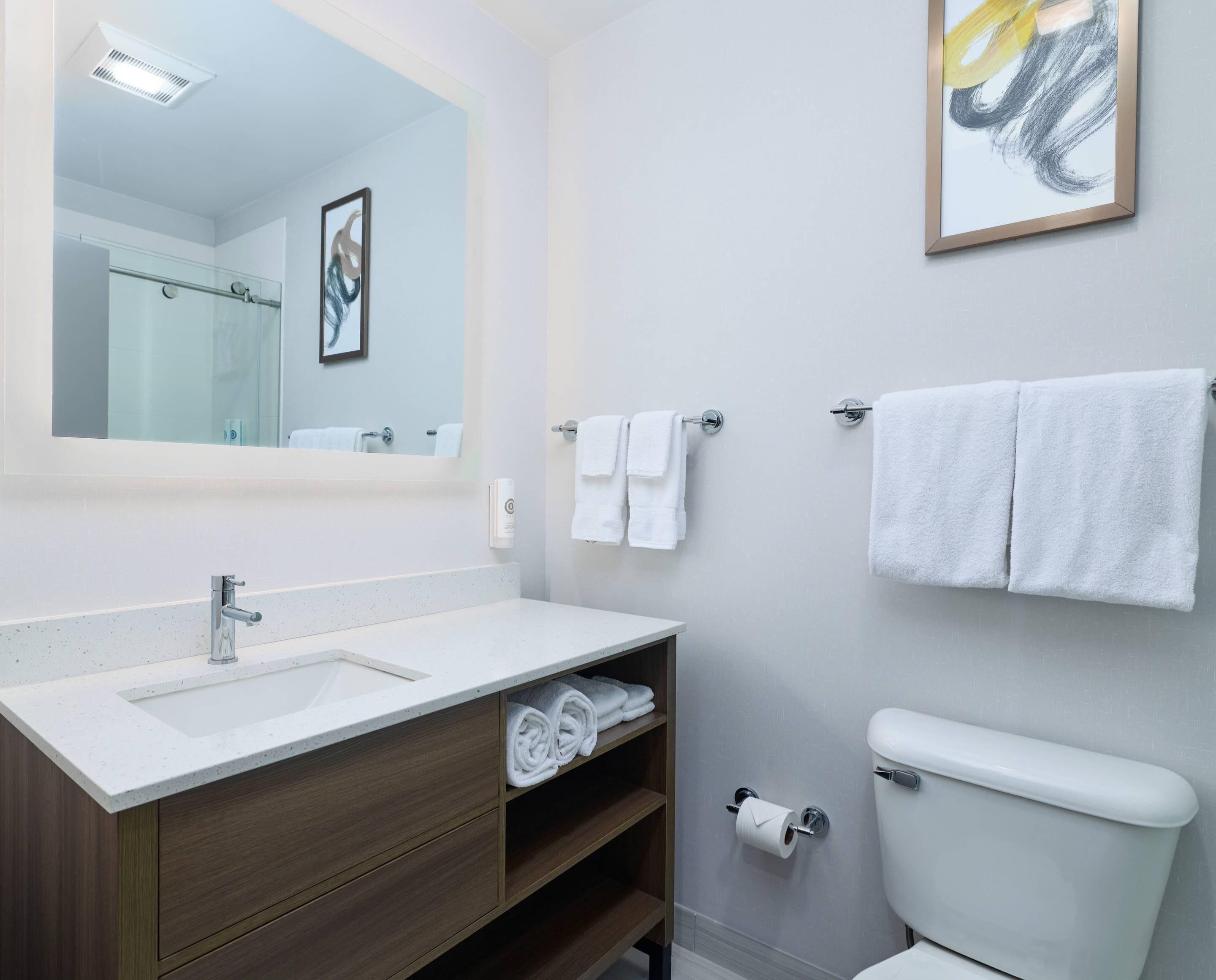 Et moderne badeværelse med en hvid vask, underskab og toilet. Der er håndklæder hængende på væggen og et indrammet billede over spejlet.