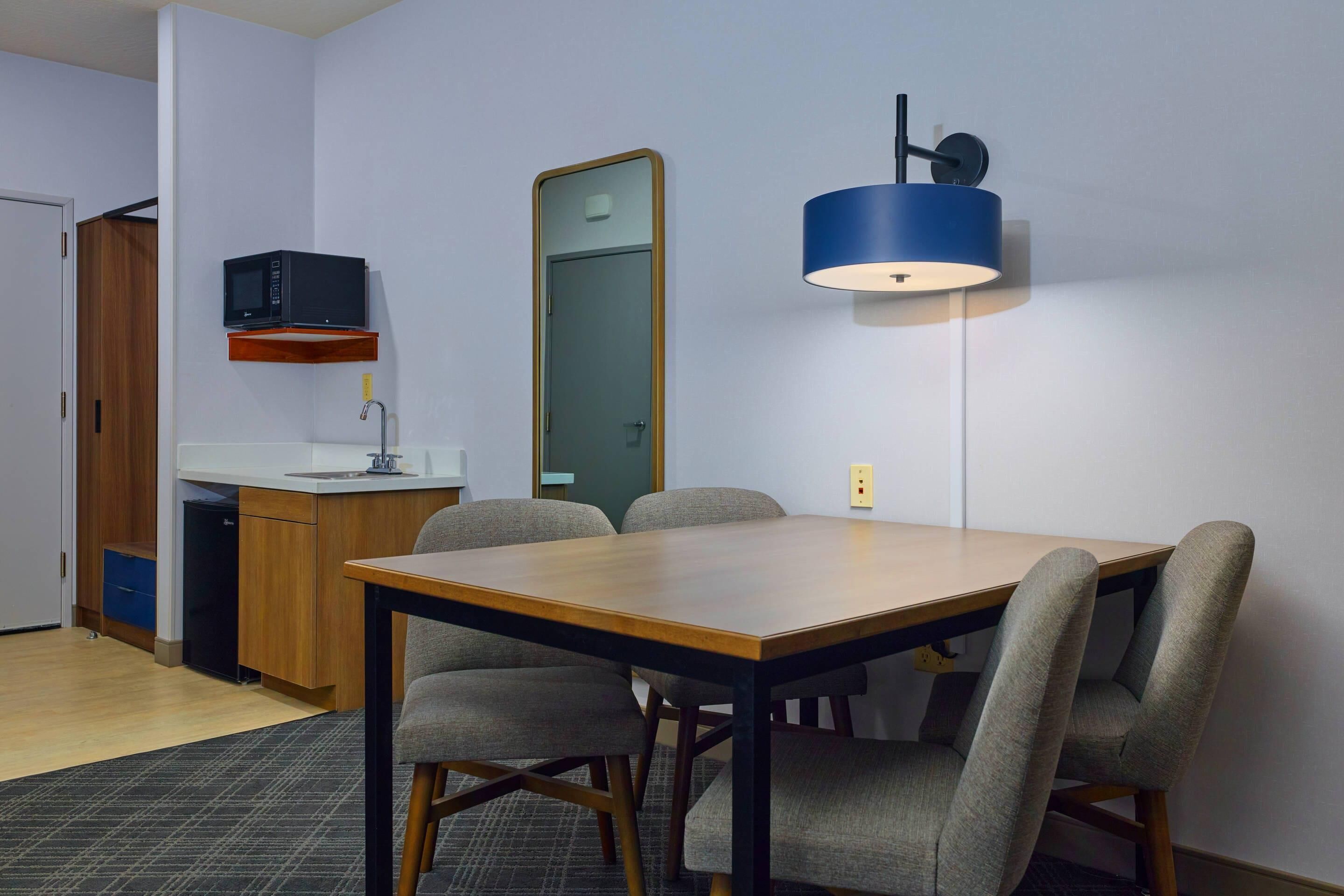 A compact kitchen with a microwave, a sink, and a wooden cabinet. There is a dining table with four chairs and a blue pendant light.