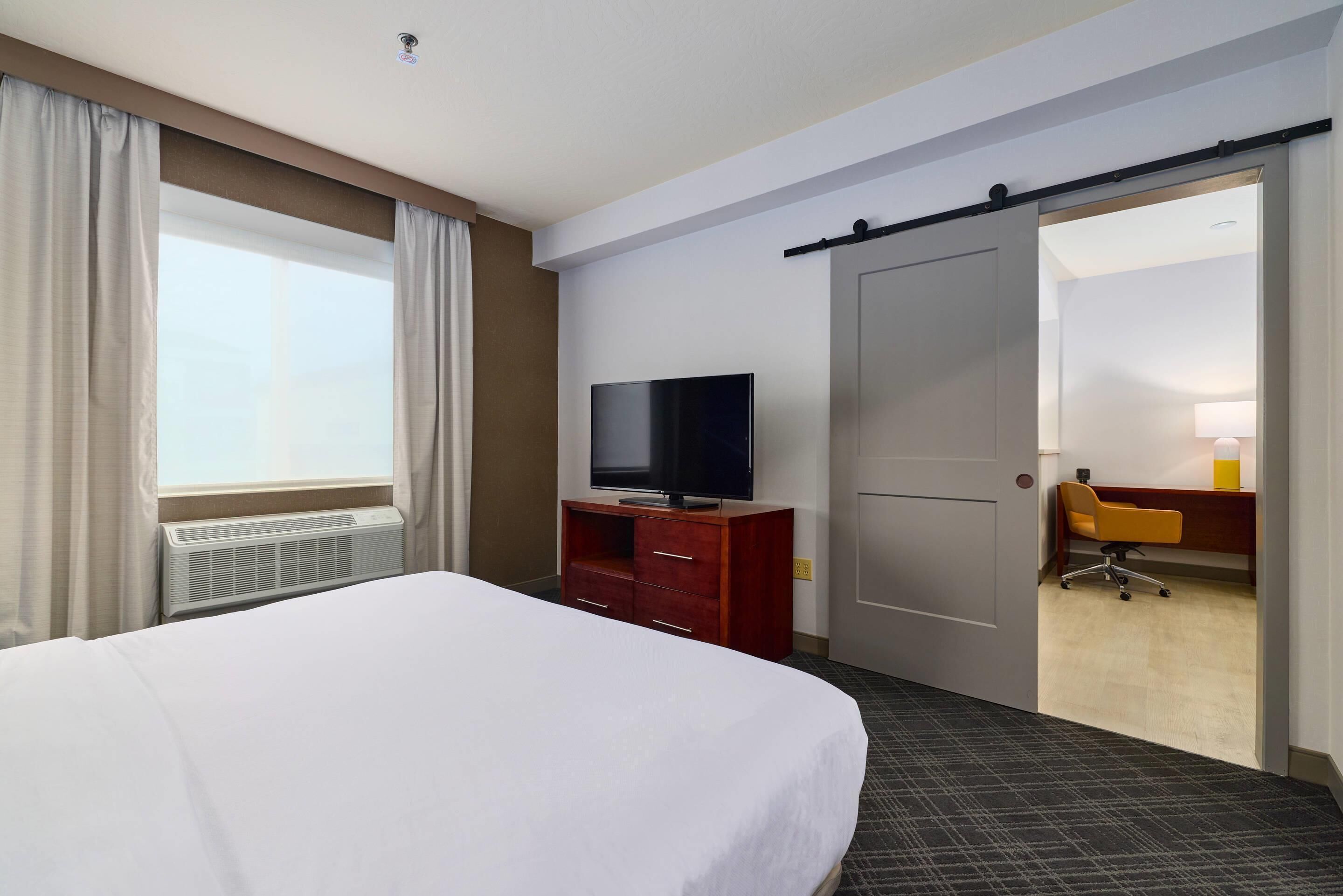 A hotel room with a bed, a TV on a wooden cabinet, a desk with a chair, and a bathroom visible through an open door.
