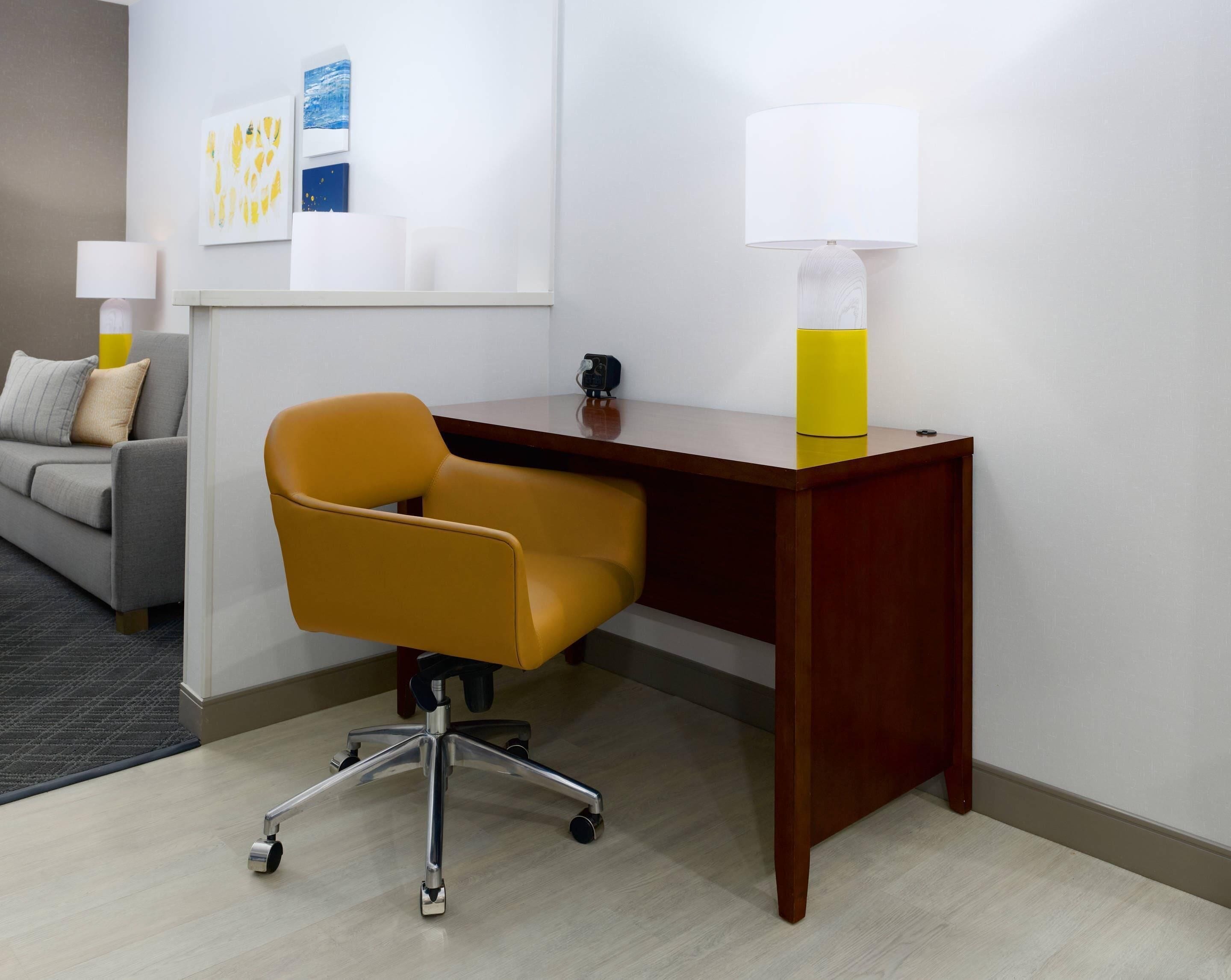 Un espacio de oficina moderno con un escritorio de madera, una silla de oficina amarilla, una lámpara de escritorio blanca y un sofá gris.